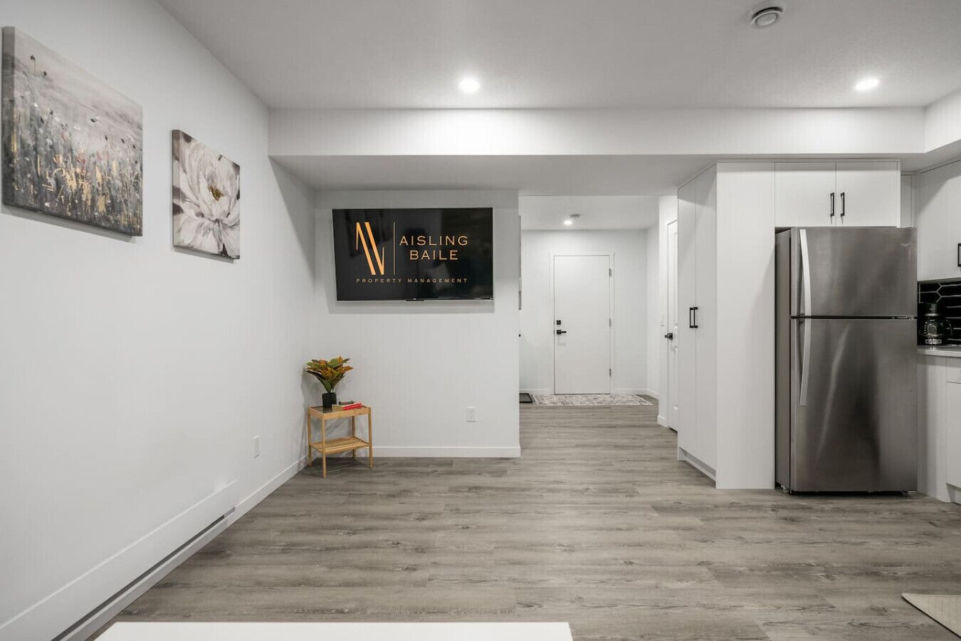 A modern kitchen with a stainless steel refrigerator, wooden flooring, and a hallway leading to a door.