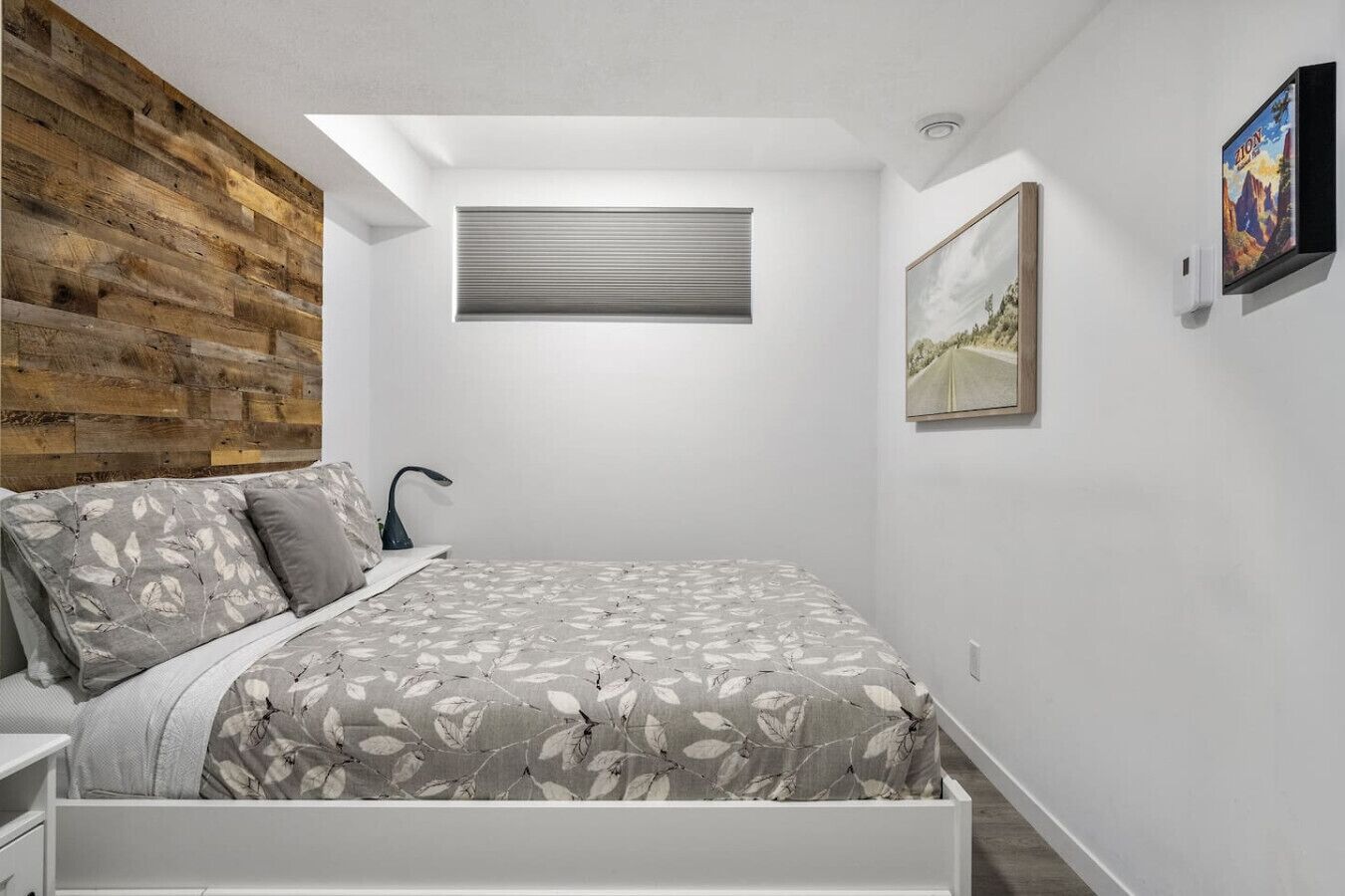 A bedroom with a bed, a nightstand, a wooden accent wall, a window with blinds, and a framed picture on the wall.