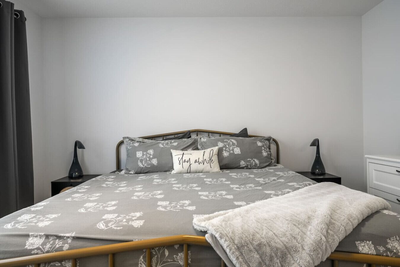 A bedroom with a bed, two bedside tables, a nightstand, a blanket, and a pillow with the text "stay awhile".