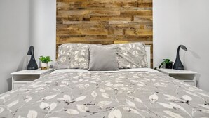 Un dormitorio con cabecera de madera, cama con cubrecama estampado con hojas, dos mesitas de noche con lámparas y una pequeña planta en maceta sobre cada una.