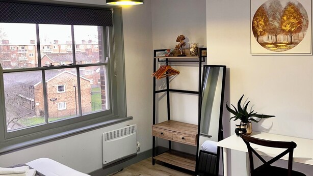 A modern bedroom with a bed, a desk, a chair, a radiator, a window with a view of buildings, and a hanging light fixture. - Stunning Ensuite Studio at Wolf’s House (London)