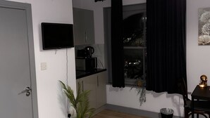 Una stanza piccola e luminosa con pavimento in legno, una TV a schermo piatto montata al muro, un angolo cottura con forno a microonde e macchina del caffè, una pianta in vaso e una finestra con tende nere.
