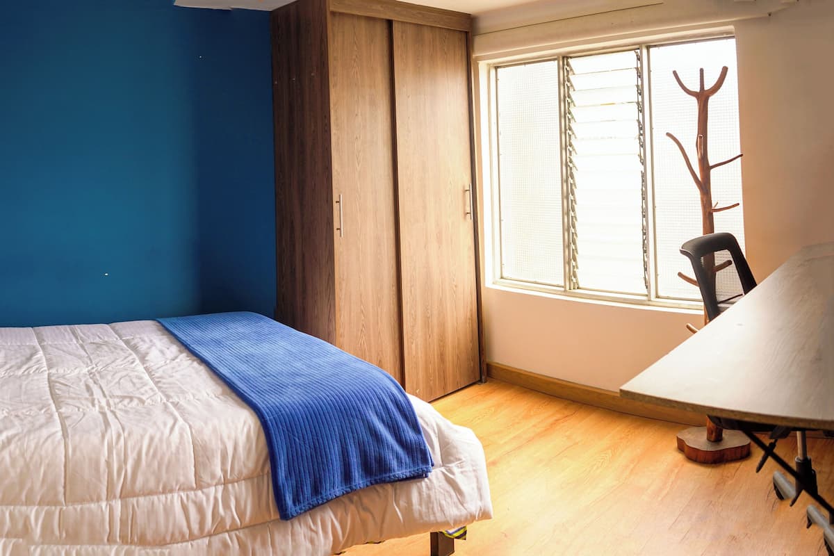 A bedroom with a bed, a blue wall, a wooden wardrobe, a window with blinds, and a chair.