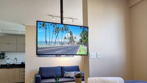 A modern hotel room with a large flat-screen TV mounted on the wall displaying a scenic view, a neatly made bed with a blue blanket, a small potted plant, and a blue sofa with a cushion. - Lindo Estúdio com Jacuzzi, Sauna e Coworking (Maceió)