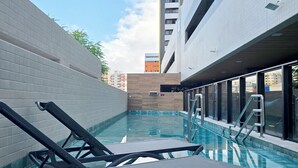 A swimming pool with lounge chairs, a tiled deck, and a view of adjacent buildings. - Lindo Estúdio com Jacuzzi, Sauna e Coworking (Maceió)