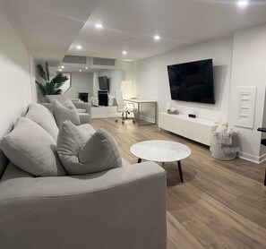 Ein modernes Wohnzimmer mit einer Eckcouch, einem runden Couchtisch, einem an der Wand montierten Fernseher und einem kleinen Beistelltisch.