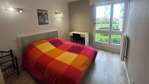 Une chambre à coucher comprenant un lit, un bureau et une fenêtre donnant sur un immeuble et de la verdure.
