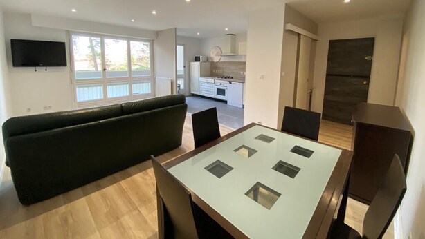 Un salon moderne avec une table à manger à plateau de verre, un canapé noir et un téléviseur fixé au mur.