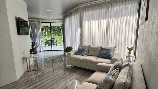 A modern living room with a beige sectional sofa, a flat-screen TV mounted on the wall, and a large window with sheer curtains. - Hidden River Lodge (La Fortuna)
