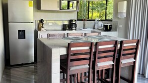 A modern kitchen with a central island, wooden chairs, a refrigerator, and a window with a view of greenery. - Hidden River Lodge (La Fortuna)