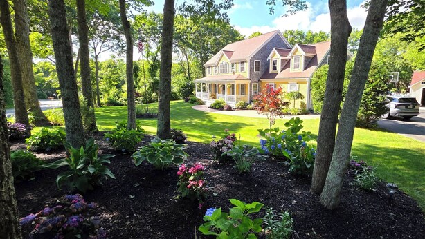 Une maison à deux étages avec un porche, entourée d’un jardin bien entretenu et d’arbres.