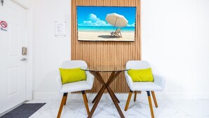 Een kamer met een witte deur, een rookvrij bordje, een lijst met een afbeelding van een strand, twee witte stoelen met gele kussens, en een ronde tafel met houten poten.