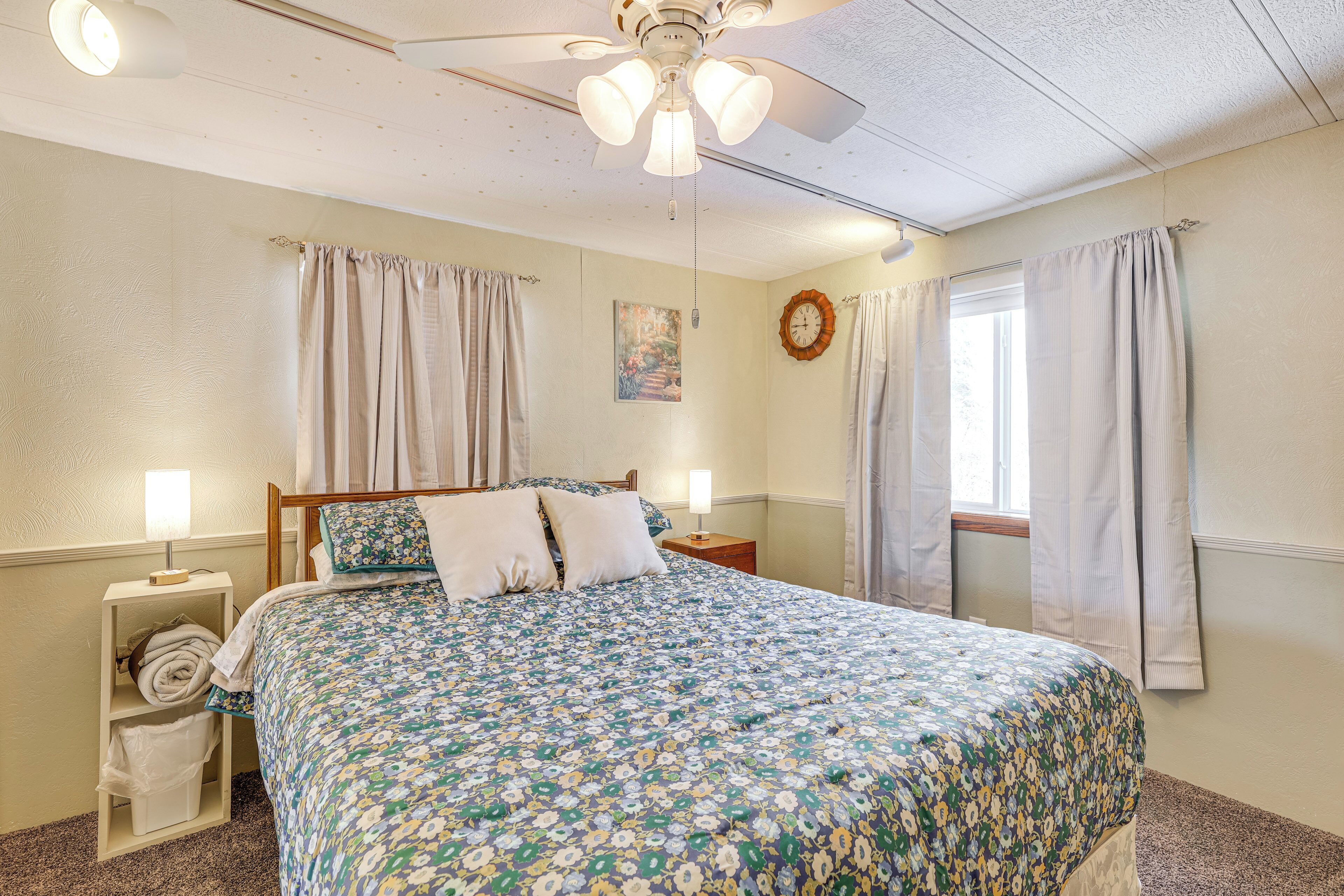 A bedroom with a floral bedspread, a ceiling fan, a nightstand with a lamp, and a window with curtains.