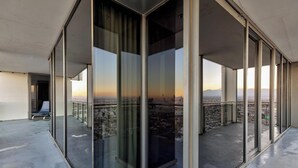 Ein modernes Hotelzimmer mit großen, raumhohen Fenstern, einem Balkon mit Blick auf die Stadt und einer Glaswand.