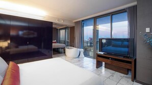Ein modernes Hotelzimmer mit einem großen Bett, einer Sitzecke mit einem weißen Sofa und einem runden Tisch, einem Flachbildfernseher und einem großen Fenster mit Blick auf die Stadt.