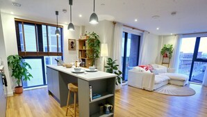 A modern apartment with a kitchen, dining area, and living room. It features wooden flooring, a large window, and a view of the city. - Northern Qtr Corner Flat With Balcony & City Views - Pass the Keys (Manchester)