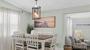 A dining area with a wooden table and white chairs, a hanging light fixture, and a painting on the wall. - Near USF, Ybor City, Busch Gardens/RayJ Stadium + TV Backyard  & Games, Slps 8! (Tampa)