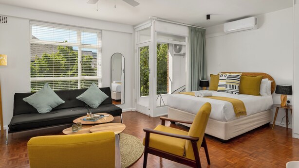 A modern hotel room with a bed, sofa, chairs, a ceiling fan, and a large window with blinds. - Vibrant Cascades Studio | Balcony | WiFi (Cape Town)