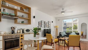A modern kitchen with wooden shelves, a dining area with white chairs, a yellow sofa, and a TV. - Vibrant Cascades Studio | Balcony | WiFi (Cape Town)