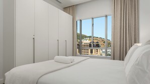 Une chambre d’hôtel moderne dotée d’un grand lit, d’une armoire et d’une fenêtre donnant sur des bâtiments et des collines.