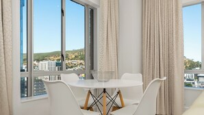 Un coin repas avec une table et des chaises blanches, une fenêtre donnant sur des bâtiments et des collines, et des rideaux de couleur beige.