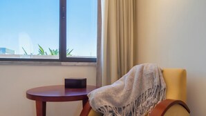 Una habitación con piso de madera, un sillón beige con una manta, una mesita de madera, una ventana con cortinas y un accesorio de iluminación fijado a la pared.