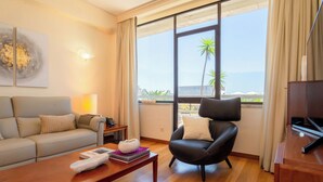 Una sala de estar moderna con un sofá, un sillón negro, una mesa de centro de madera y un ventanal con vistas al mar y palmeras.