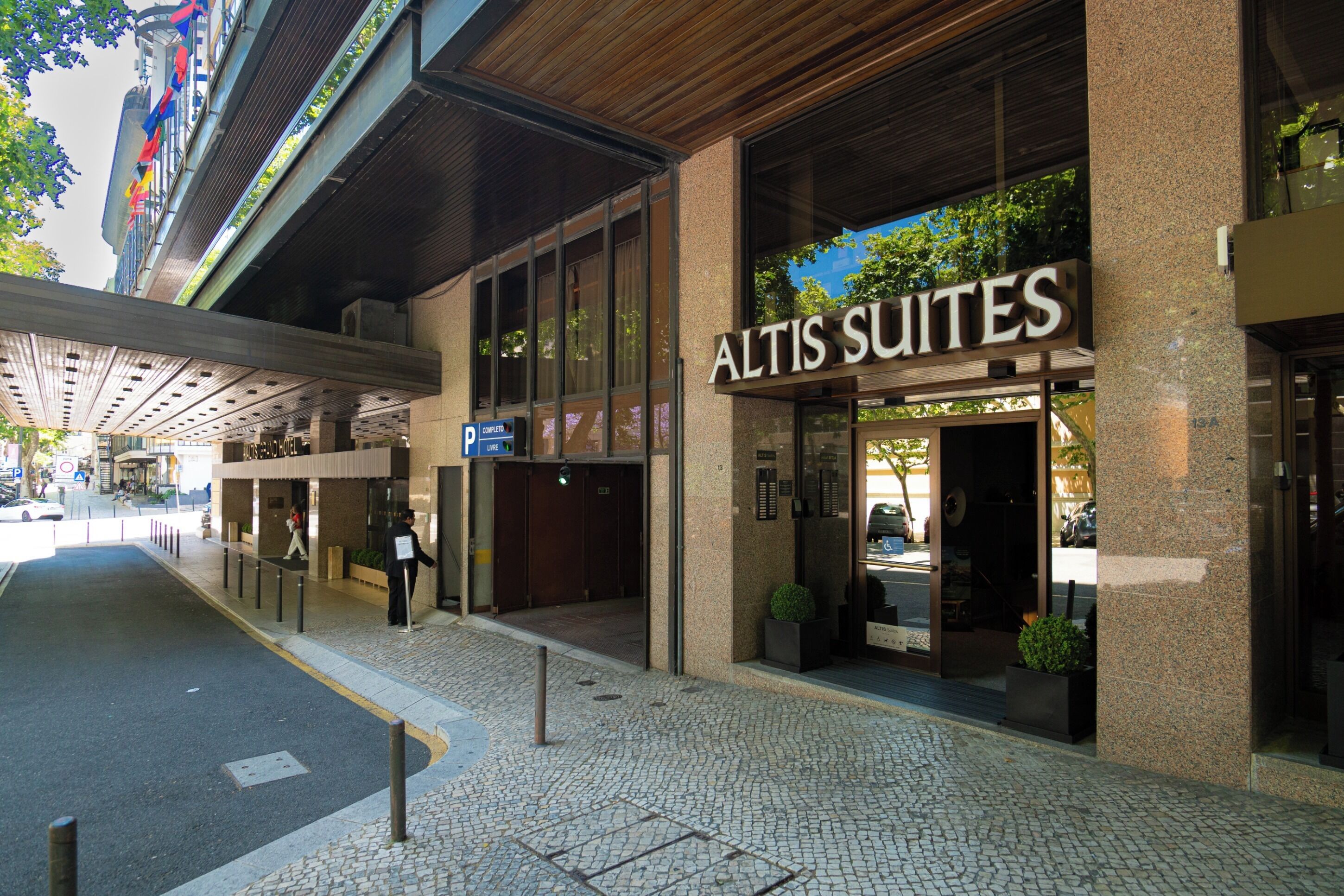 Exterior do Altis Suites, com fachada moderna e grandes janelas de vidro, e entrada destacada.