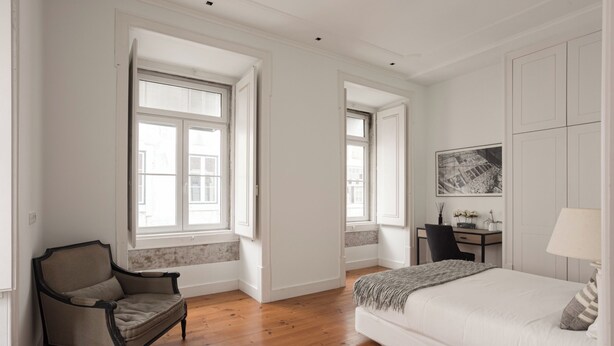 Une chambre avec un grand lit, une chaise, un bureau avec une lampe et deux fenêtres.