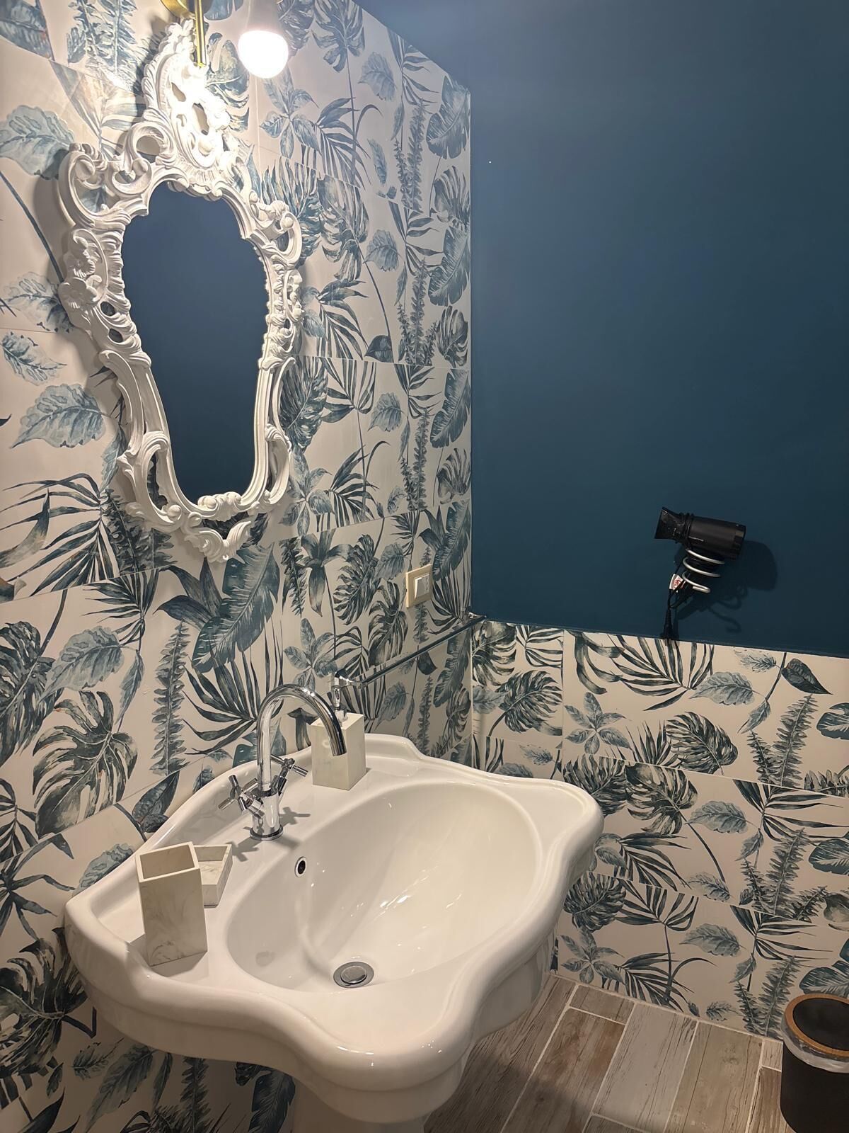 A bathroom with a white sink, a mirror with an ornate frame, and a blue wall with tropical leaf wallpaper.