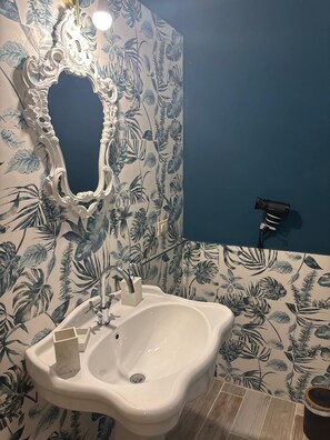 Une salle de bain avec un lavabo blanc, un miroir doté d’un cadre orné et un mur bleu recouvert d’un papier peint à motifs tropicaux.