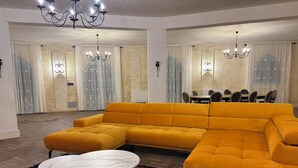 A modern living room with a large yellow sectional sofa, two round marble tables, a patterned rug, and a dining area with a table and chairs.