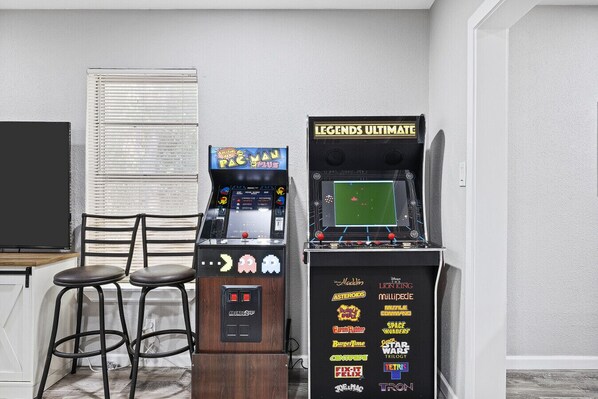 Två arkadspel, varav det ena är märkt med "LEGENDS ULTIMATE" och det andra med "PAC-MAN PLUS", står i ett rum med en vit vägg, ett fönster med persienner och en trästol.