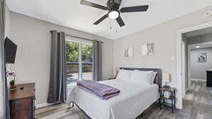A bedroom with a bed, a ceiling fan, a nightstand, a window with curtains, and a view of the outdoors. - Waco Oasis | 5 min to downtown, Magnolia, & Baylor (Waco)
