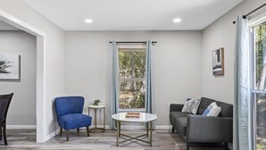 A modern living room with a blue armchair, a grey sofa, a small round table, and a window with curtains. - Waco Oasis | 5 min to downtown, Magnolia, & Baylor (Waco)