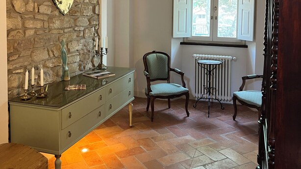Une pièce avec un mur en pierre, un panneau en forme de cœur, une commode verte, deux chaises, un radiateur et une fenêtre avec des volets.