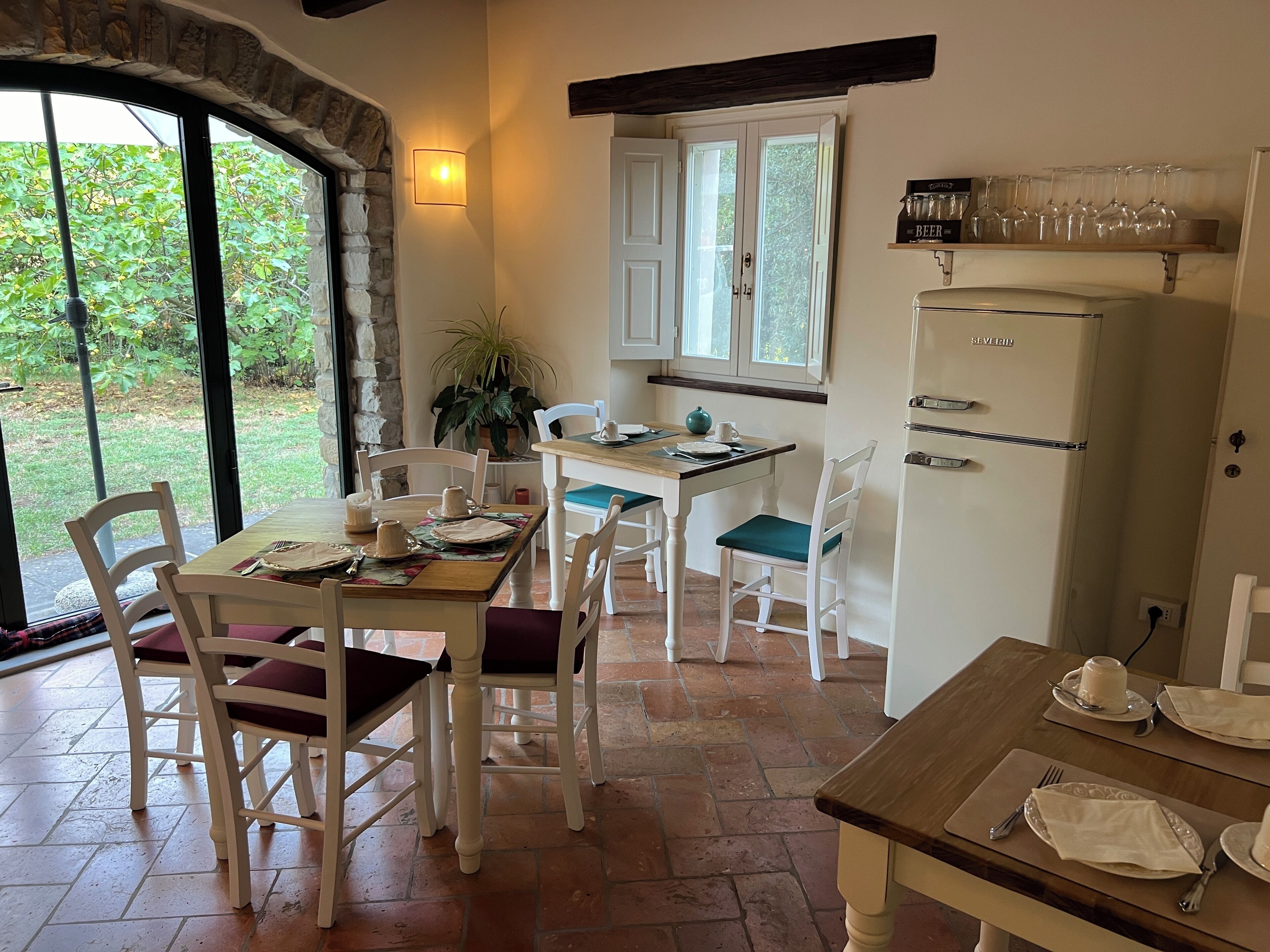 A dining area with a table set for four, a refrigerator, and a window with a view of greenery.