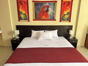 A hotel room with a large bed, two bedside tables with lamps, and three paintings on the wall. - DORAL INN HOTEL (Iquitos)