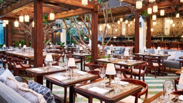 An outdoor dining area with wooden tables and chairs, string lights, and a patterned wall in the background.