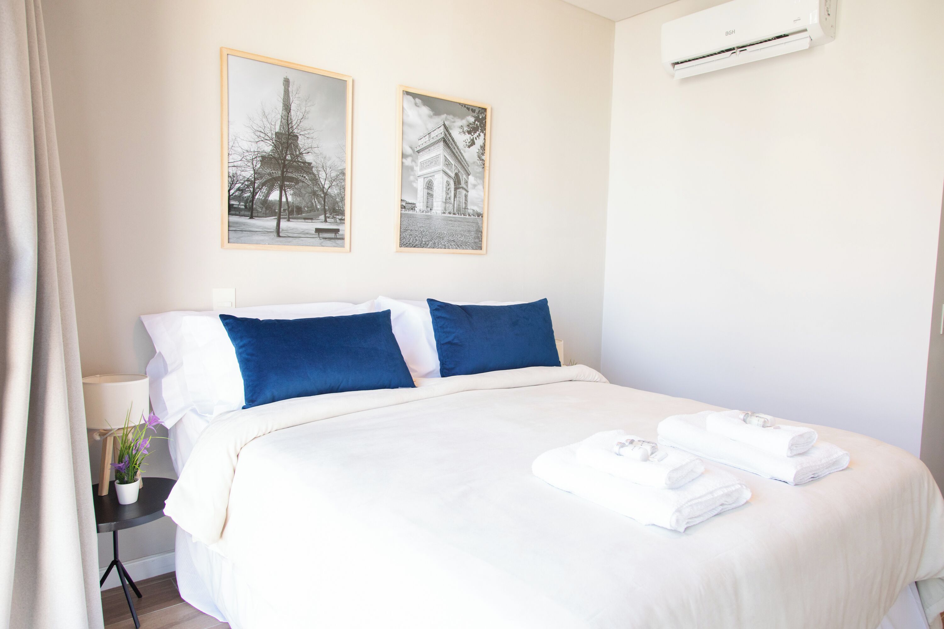 Een hotelkamer met een bed, witte beddengoed, blauwe kussens, een tafeltje met een lamp en een bloempot, en twee ingelijste foto’s aan de muur.