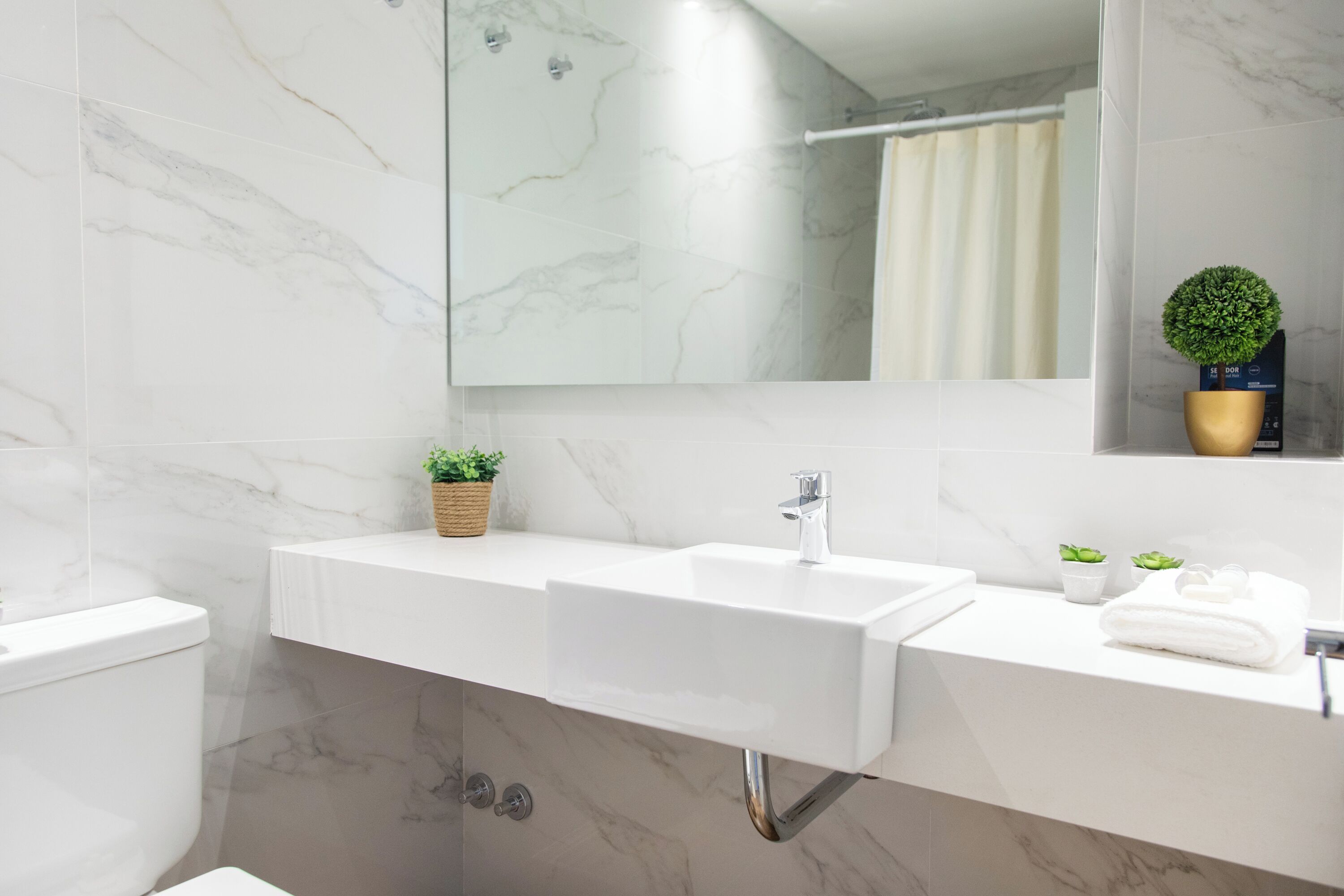Une salle de bain moderne avec un lavabo blanc, un miroir et des toilettes. De petites plantes en pot sont posées sur le comptoir du lavabo.