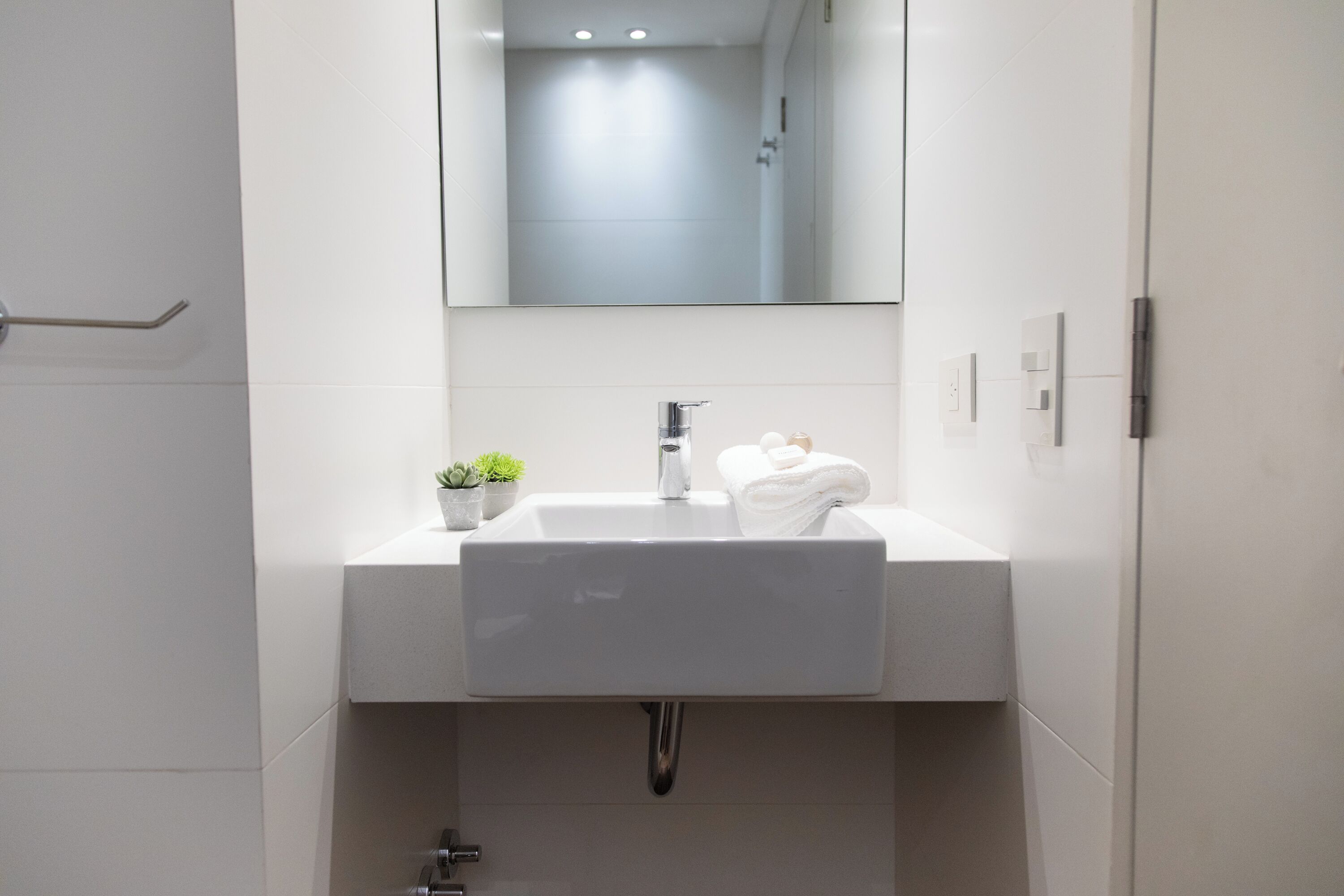 Un bagno moderno con un lavandino bianco, uno specchio sopra e una piccola pianta in vaso sul ripiano.