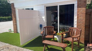 Un patio con dos sillas de madera y un banco, una planta en maceta y una puerta corredera blanca.