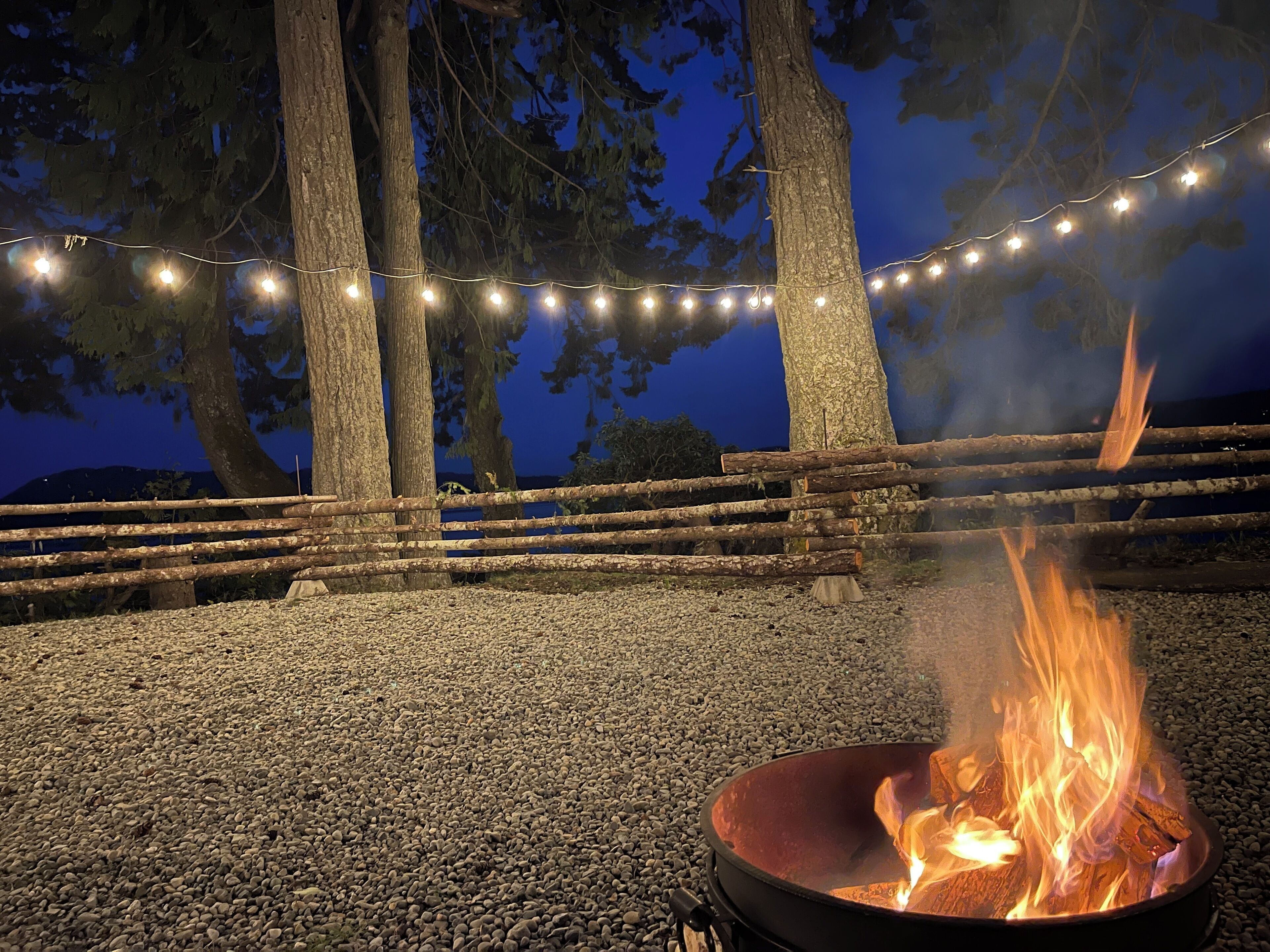 Ein Lagerfeuer mit Metall-Feuerstelle, umgeben von Kies, unter Lichterketten, mit Bäumen und einem Holzzaun im Hintergrund.