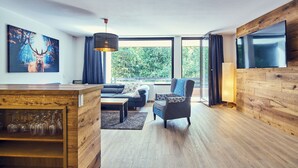 A modern living room with a wooden floor, a sofa, a patterned armchair, a coffee table, and a large TV mounted on the wall.