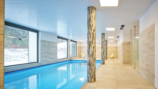 A modern indoor swimming pool with clear windows, wooden flooring, and a minimalist design.