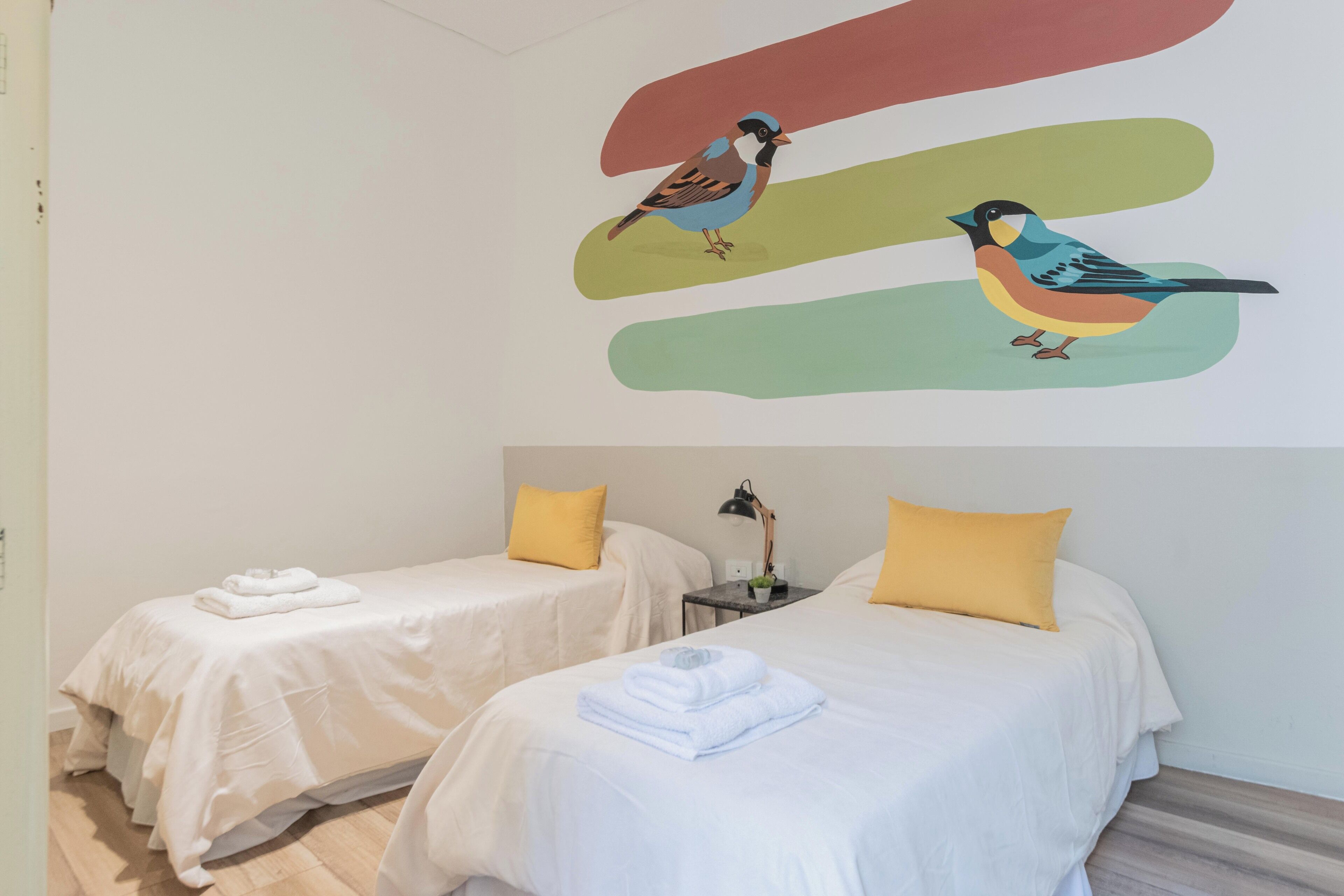 Quarto de hotel com duas camas, cada uma com um cobre-leito branco e um travesseiro amarelo. Há uma obra de arte na parede com duas aves e formas abstratas.