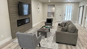 A modern living room with a flat-screen TV mounted on a wooden wall, a grey armchair, a grey sofa, and a patterned rug. - Modern and cozy home on Lakeview (Lake Oswego)