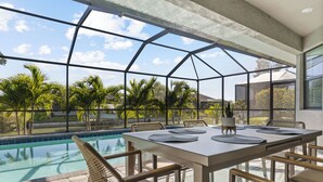 Zona de comedor junto a la piscina, con cerramiento de vidrio, asientos exteriores y vistas a la piscina y palmeras.