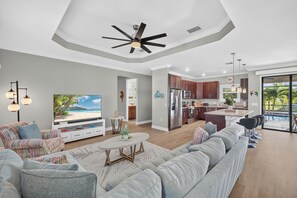 Una sala de estar moderna con un televisor grande, un ventilador de techo y una zona de comedor con una cocina al fondo.